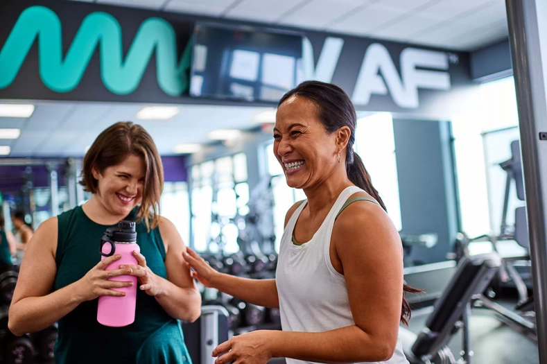 Two women smiling and talking inside a gym near cardio equipment